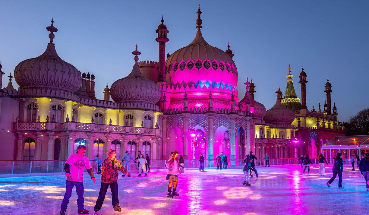 Image shows skaters at the Royal Pavilion ice rink, skating in front of the palace which is light in a warm pink glow. Image shows skaters at the Royal Pavilion ice rink, skating in front of the palace which is light in a warm pink glow.