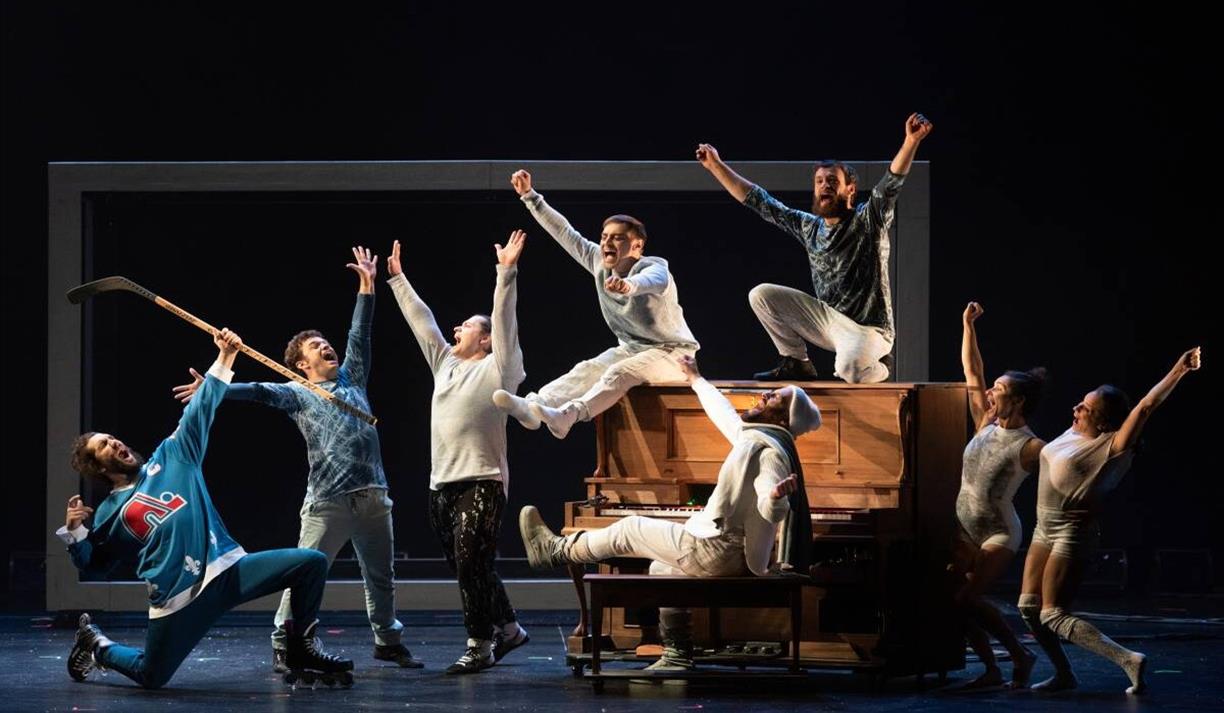Actors on stage in Blizzard at Brighton Dome. Image shows actors in pale and blue outfits sitting on and around a piano on stage. They have their hand Actors on stage in Blizzard at Brighton Dome. Image shows actors in pale and blue outfits sitting on and around a piano on stage. They have their hand
