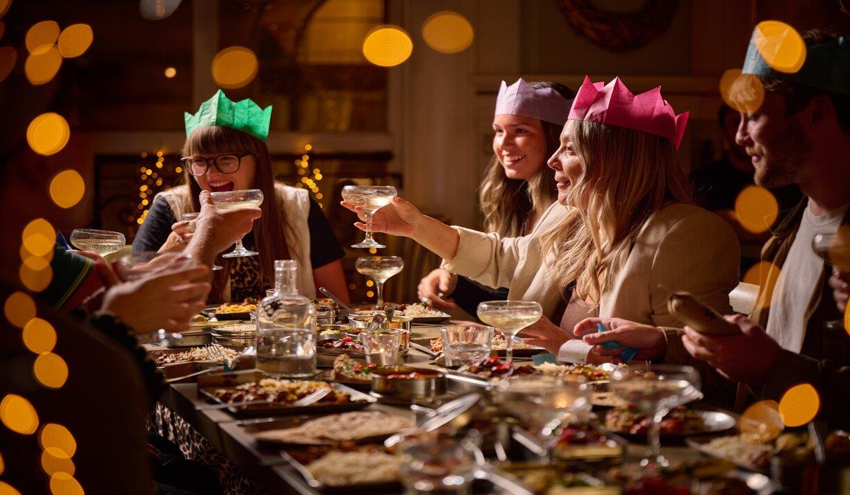Partygoers enjoying a meal at Mowgli Brighton. a group of friends wearing colourful Christmas hats raise a toast, there are dishes of food in front of Partygoers enjoying a meal at Mowgli Brighton. a group of friends wearing colourful Christmas hats raise a toast, there are dishes of food in front of