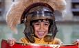 A young boy wearing a Roman helmet standing behind a shield by The Great Bath at The Roman Baths