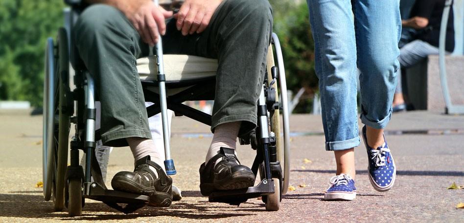 A close up of the legs of a person seated in a wheelchair with the legs of a person walking alongside.