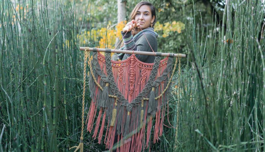 A woman standing in tall grass holding a macrame tapestry