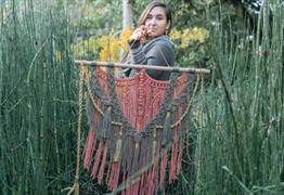 A woman standing in tall grass holding a macrame tapestry