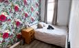Double bed with bedside drawers and bright floral wallpaper. Bed is positioned next to window.