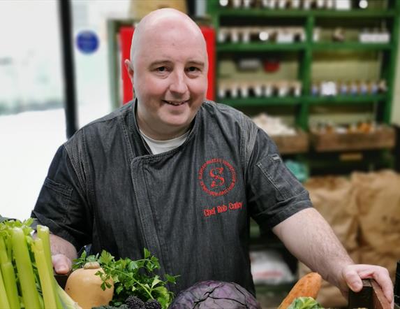 a man stands with a box filled with fresh vegetables including carrots, parsnips, celery, onions and cabbage