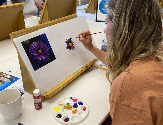 A woman painting a purple flower using a reference image attached to her canvas.