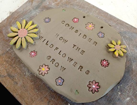 A ceramic sign with flowers carved into and embellishing it. It reads "Consider how the Wild Flowers grow"
