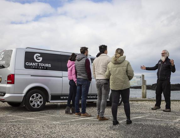 tour guide, Flip, talks to a group of tourists standing beside a van branded with the 'Giant Tours' logo