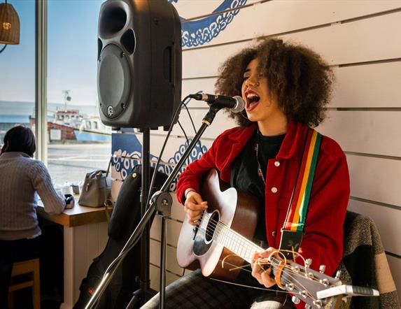 Image shows a lady singing and playing guitar.  In the background there are two people sitting looking out the window at the harbour in Portstewart.