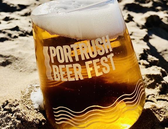 A glass of frothing beer on the sand. Text on the glass reads "Portrush Beer Fest"