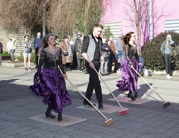 Irish dancers entertaining the crowds in Drumceatt Square, dressed in purple and black dresses with brooms in hand.