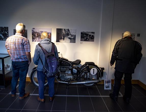 Images shows three men looking at old photographs on the wall with a vintage bike on display in front of them