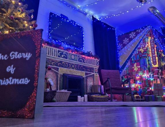 a quiet living room scene is depicted lit up with Christmas lights and a tree. a book is propped up on the floor and the front cover reads 'The Story