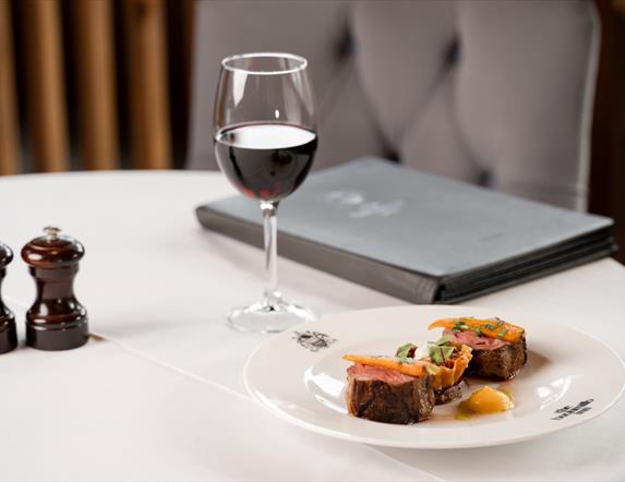 Image shows a chair and dining table covered in a white cloth, with a plate of food, a glass of red wine, a menu and salt/pepper grinders