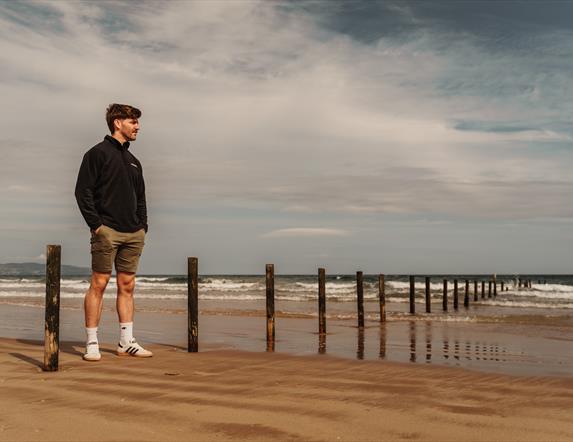 Artist Ruairi Mooney stands on a beach with the ocean in the background