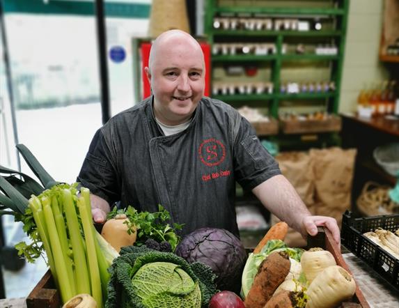 a man stands with a box filled with fresh vegetables including carrots, parsnips, celery, onions and cabbage