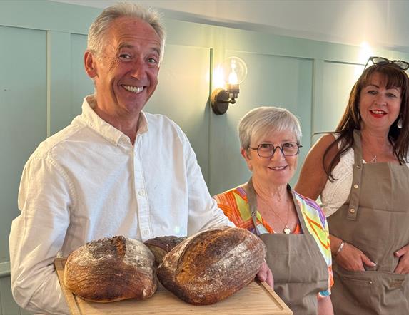 A man stands smiling holding loaves of fresh sourdough bread. Two women in aprons stand behind him and to the right.