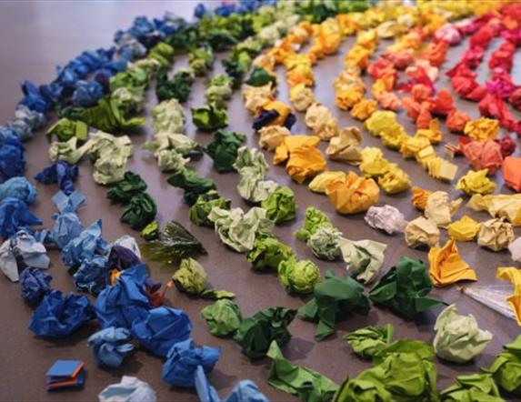 A number of crumpled pieces of coloured paper form a larger rainbow, in a circular shape.