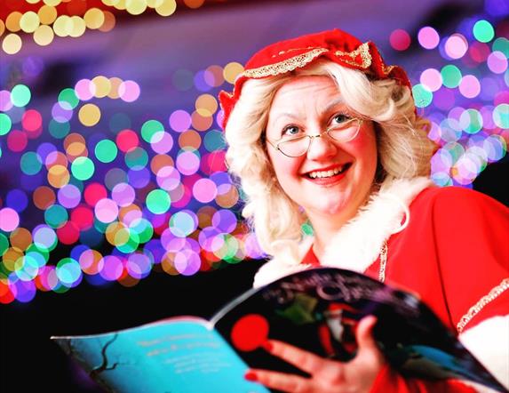 smiling lady dressed in red with small glasses standing in front of a blurry sparkly lights background.