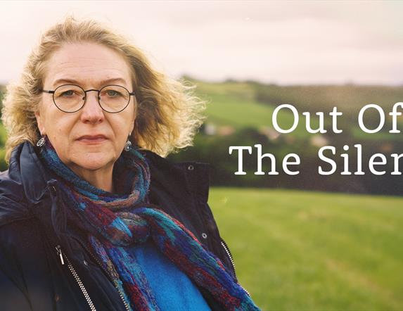 A woman with blond hair and round spectacles stands in a field looking towards the camera. Beside her the title "Out of the Silence" is written in whi