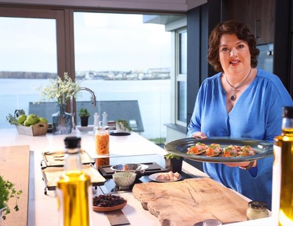 Paula McIntyre stands at a kitchen island which overlooks the sea. she is holiday a tray of food