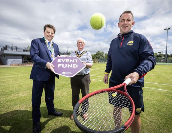 2 men hold a sign saying 'Event Fund' while another bounces a tennis ball on a racket