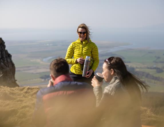 a man and woman sitting on a cliff drinking from cups. a woman stands in front of them with a thermal flask