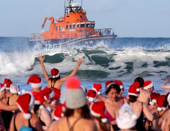 people in santa hats and swimwear watch on as a RNLI lifeboat cruises on the waves