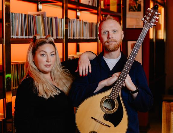 Image of Aideen McGinn and Martin Coyle tanding in from of a bookcase.  Martin is holding a guitar.