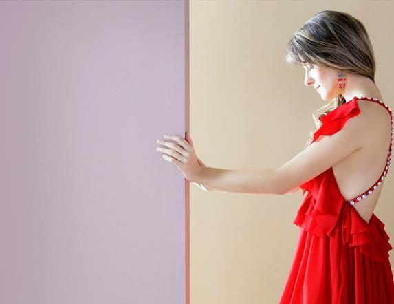 Image shows a woman wearing a red dress holding her hand against a wall. The left side of the wall is purple and the right side cream.