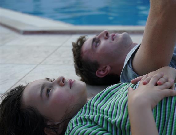 Iamge of a girl (wearing a green stripped t-shirt) and a boy (wearing a blue t-shirt) laying on their backs beside a swimming pool
