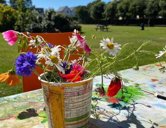 A bundle of colourful flowers potted in a makeshift planter, with a park and clear blue sky visible in the background.