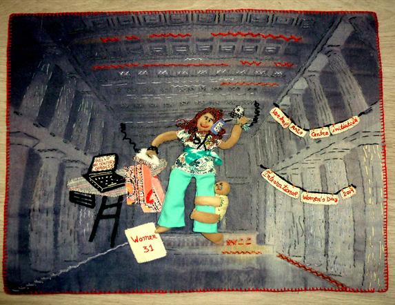 Image shows a scene with an arpillera doll with a young baby hanging on to her leg and a laptop on a desk