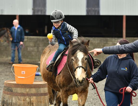 a child aims a ball towards a bucket while on horseback as Crindle Stables' instructors lead the horse