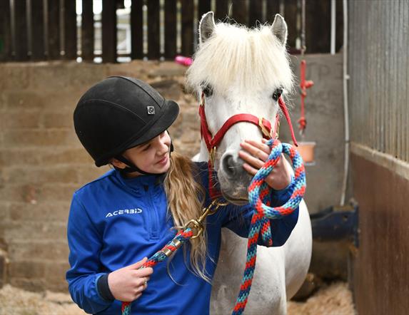 Image shows a young girl in a blue top and black riding helmet petting a white horse