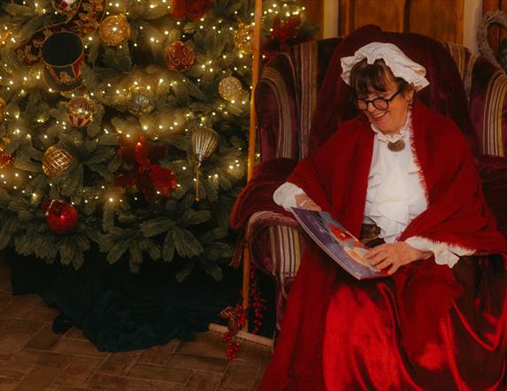 Mrs Claus sits in an armchair by the Christmas tree