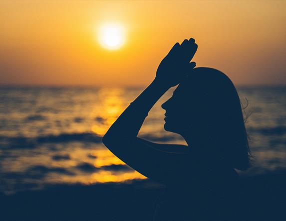 a person raises joined hands to their forehead with the sunset and the sea in the background