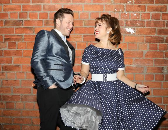 Picture of a man and a woman smiling at each other leading up against a wall. She is wearing a navy and white polka dot dress with a wide white belt,