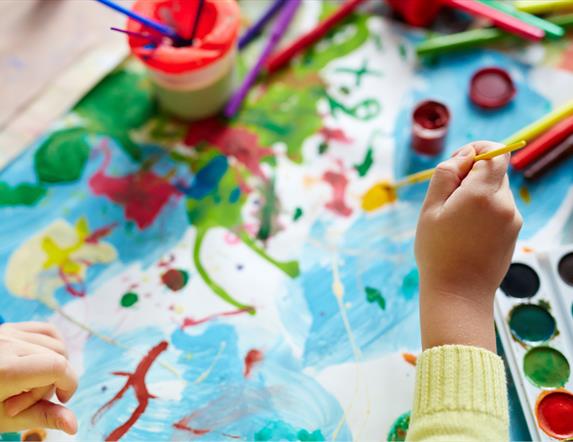 Child's hands paint across a messy canvas, surrounded by various art supplies.