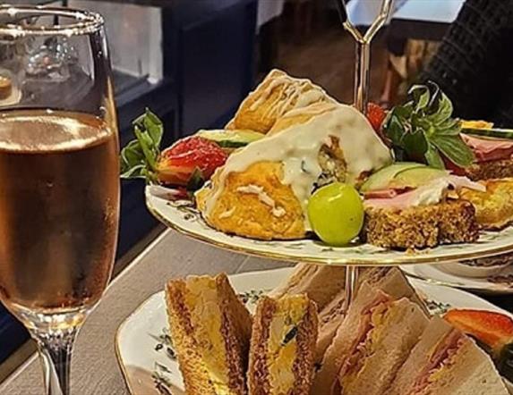 glass of prosecco next to a tiered cake stand holding sandwiches, fruit and scones