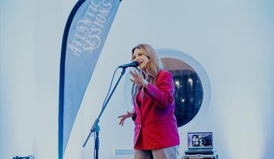 a woman sings in to a microphone on stage with a branded Atlantic Sessions flag beside her