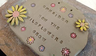 A ceramic sign with flowers carved into and embellishing it. It reads "Consider how the Wild Flowers grow"