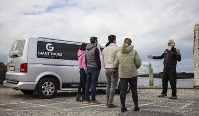 tour guide, Flip, talks to a group of tourists standing beside a van branded with the 'Giant Tours' logo