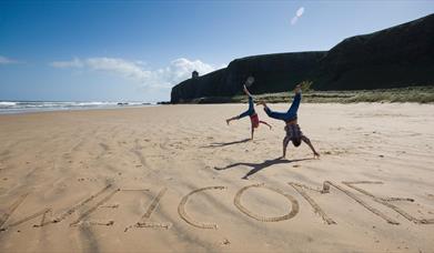 Image of beach with cliffs and sea in the background. The iconic Mussenden Temple is visible in the distance. In the foreground, 'welcome' has been ca