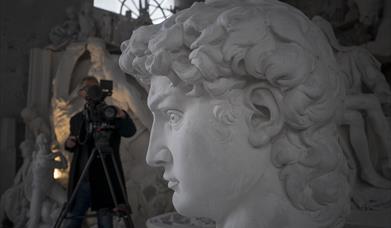 close up image of a sculpted head with a videographer standing in the background with a camera on a tripod pointed towards it