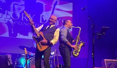 musicians from the Glory Days Bruce Springsteen Tribute band perform on stage. One is strumming a guitar while the other plays a saxophone, and a drum