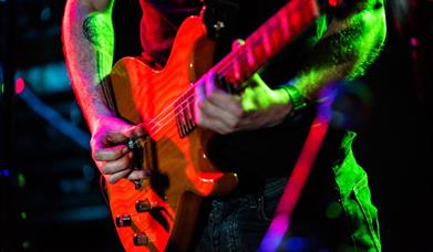 Close up image of an electric guitar being played. The guitarist has a tattoo on his arms and is wearing dark jean and a dark coloured t shirt.