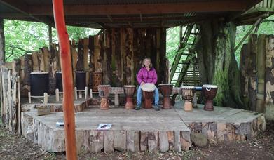 a person sitting outdoors on a wooden stage with a selection of  African Djembe drums sitting beside them.