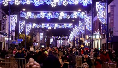 Image shows Christmas lights shining above a busy street with people shopping