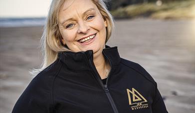 Claire O'Kane of Mussenden Sea Salt stands smiling beneath Mussenden Temple, wearing a black zip up fleece with her company logo in gold.